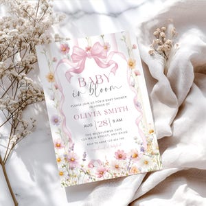 May include: A baby shower invitation with the text "Baby in Bloom". The design features a pink bow, ribbons, and floral accents. The invitation includes details for Olivia Smith's baby shower, including the date, time, and location.
