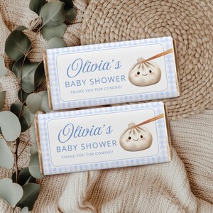 May include: Two chocolate bars with light blue gingham wrappers. Each wrapper has a white label with the text "Olivia's BABY SHOWER THANK YOU FOR COMING!" and a cartoon dumpling illustration. The bars are on a beige knitted surface.