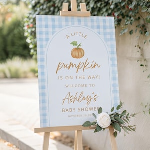 May include: A wooden easel displays a baby shower welcome sign. The sign has a light blue gingham border and features the text "A Little Pumpkin is on the Way!" with a pumpkin illustration. The sign welcomes guests to Ashley's baby shower.
