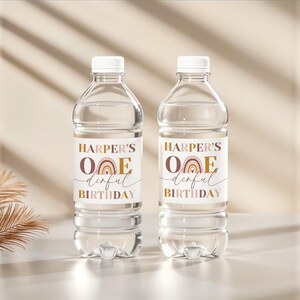Peut inclure: Deux bouteilles d'eau en plastique transparent avec des bouchons blancs. Chaque bouteille porte une &eacute;tiquette avec le texte "HARPER'S ONE derful BIRTHDAY" et un arc-en-ciel marron et dor&eacute;. Les bouteilles sont sur une surface claire.