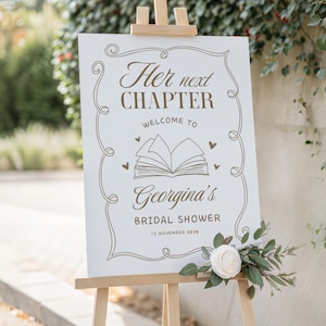 May include: A white sign on a wooden easel with the text "Her next chapter, Welcome to Georgina's Bridal Shower, 12 November 2030" in brown script. The sign is decorated with a floral design and a white rose.