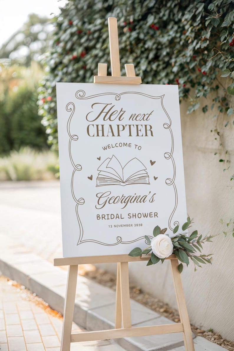 May include: A white sign on a wooden easel with the text "Her next chapter, Welcome to Georgina's Bridal Shower, 12 November 2030" in brown script. The sign is decorated with a floral design and a white rose.