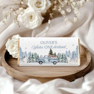 May include: A chocolate bar with a winter-themed design. The bar features a light blue pickup truck carrying a deer and a forest scene with the text "Oliver's Winter One derland". The bar is presented on a wooden tray with white roses.