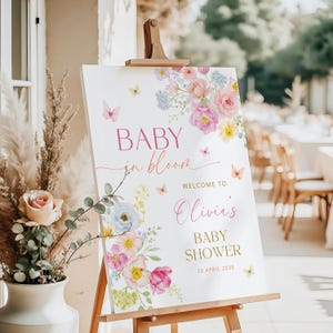 May include: A white rectangular welcome sign on a wooden easel featuring floral illustrations in pink, yellow, and blue, with butterflies and the text: BABY in bloom, WELCOME TO Olivia's BABY SHOWER, 30 APRIL 2030. The sign has a smooth, matte finish.