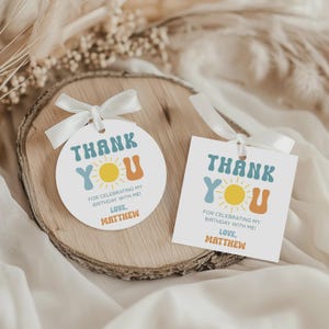 May include: Two white gift tags with a retro design. The tags have the words "THANK YOU" in blue and orange, with a yellow sun graphic. The text "FOR CELEBRATING MY BIRTHDAY WITH ME! LOVE, MATTHEW" is also included. The tags are tied with white ribbons.