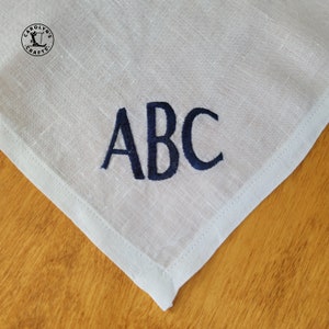 May include: White linen handkerchief with navy blue embroidered initials "ABC". The handkerchief features a stitched border and is shown on a wooden surface. The upper left corner has a logo that says "Carolyn's Crafts".