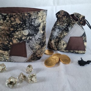Peut inclure: Deux sacs à dés en tissu avec un motif marbré noir, beige et crème, avec des patchs marron et gris. Les sacs sont accompagnés de dés transparents avec des inclusions florales et de pièces de monnaie dorées. Un sac est ouvert, l'autre est fermé par un cordon.