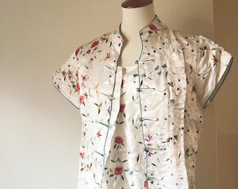 May include: A cream-colored, short-sleeved blouse and matching top with a floral pattern of red, purple, and blue flowers and green leaves. The blouse has a mandarin collar and frog closures. The edges are trimmed with green piping. The outfit is displayed on a mannequin.