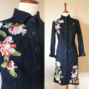May include: A dark blue denim dress with embroidered floral details. The dress has a button-down front and long sleeves.
