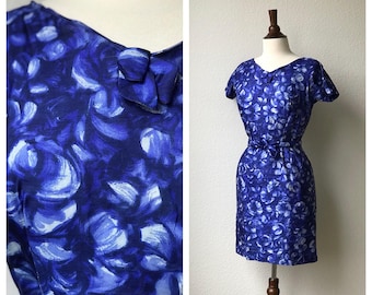 May include: A blue and white floral print dress with short sleeves and a bow at the neckline. The dress is fitted and has a skirt that falls to the knees.