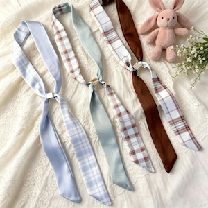 May include: A collection of satin neck scarves in various colors, including light blue, brown, and sage green. Some scarves feature a plaid pattern, while others are solid. Each scarf is connected by a silver-colored metal clasp. A pink plush bunny is in the background.