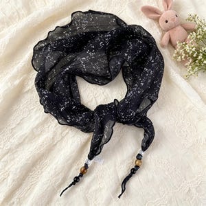 May include: A black scarf featuring a white speckled pattern and a ruffled edge. The ends of the scarf are tied and decorated with a series of beads in clear, brown, gold, and black colors, creating a distinct finish to the lightweight fabric.