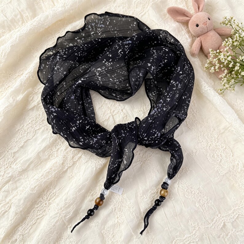 May include: A black scarf featuring a white speckled pattern and a ruffled edge. The ends of the scarf are tied and decorated with a series of beads in clear, brown, gold, and black colors, creating a distinct finish to the lightweight fabric.