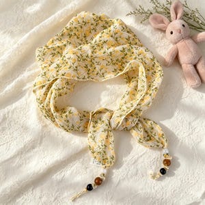 May include: A lightweight scarf featuring a yellow floral pattern on a white background. The scarf ends are decorated with dangling beads in brown, black, and white colors. A small plush pink rabbit toy is positioned near the scarf fabric.