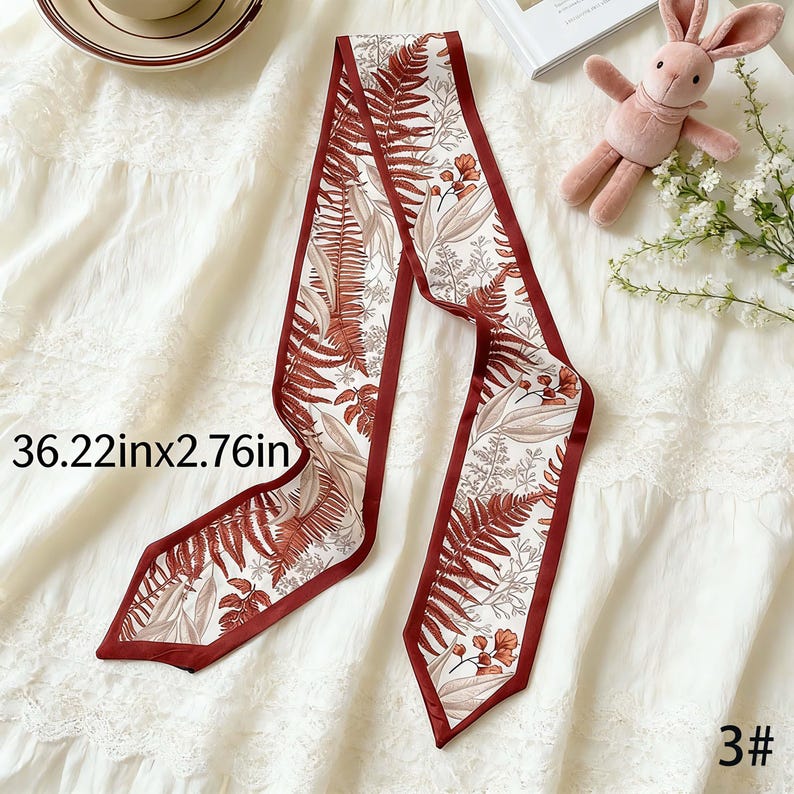 May include: A decorative scarf with a botanical print. The scarf is white with a repeating pattern of brown ferns and flowers, and has a dark red border. The scarf measures 36.22 inches long and 2.76 inches wide.