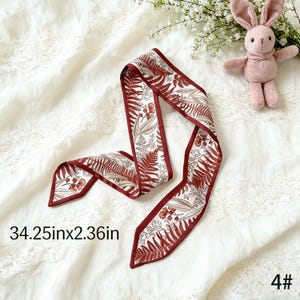 May include: A decorative scarf with a botanical print featuring ferns and flowers in shades of brown and beige, outlined with a dark red border. The scarf measures 34.25 inches long and 2.36 inches wide.