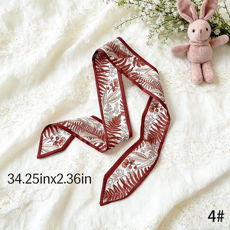 May include: A decorative scarf with a botanical print featuring ferns and flowers in shades of brown and beige, outlined with a dark red border. The scarf measures 34.25 inches long and 2.36 inches wide.
