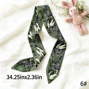 May include: A long, dark green scarf with a botanical print featuring ferns and other foliage. The scarf measures 34.25 inches long and 2.36 inches wide. The edges are a darker green, and the background is a dark navy blue.