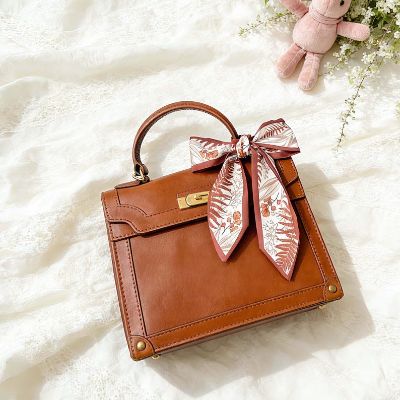 May include: A brown leather handbag with a top handle and gold-colored hardware. A decorative scarf with a floral pattern in shades of brown and white is tied to the handle. The bag is square-shaped and sits on a white lace surface.