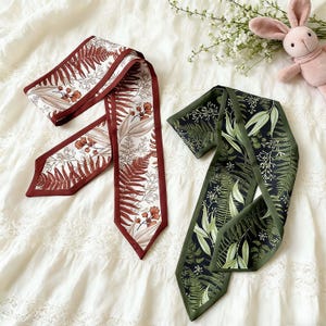 May include: Two silk scarves with botanical patterns. One scarf features a white background with brown and rust-colored fern and floral designs, framed by a dark brown border. The other scarf has a dark green background with green and white leaf patterns, framed by a dark green border.