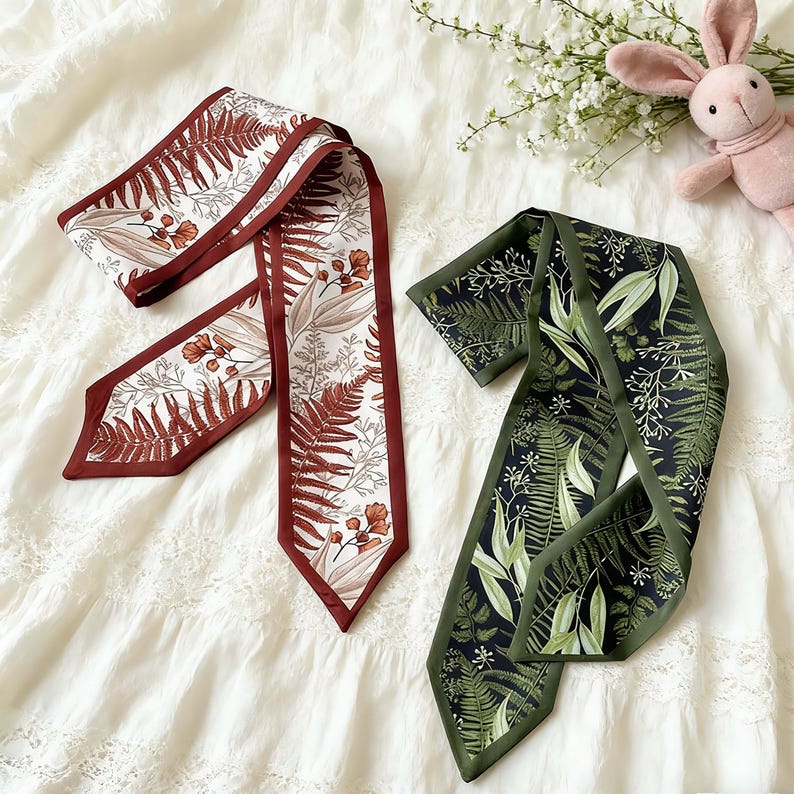 May include: Two silk scarves with botanical patterns. One scarf features a white background with brown and rust-colored fern and floral designs, framed by a dark brown border. The other scarf has a dark green background with green and white leaf patterns, framed by a dark green border.