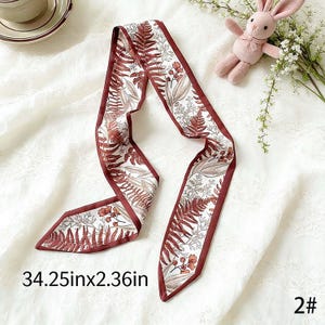 May include: A decorative scarf with a botanical print featuring ferns and flowers in shades of brown and white. The scarf has a dark red border and measures 34.25 inches long and 2.36 inches wide.