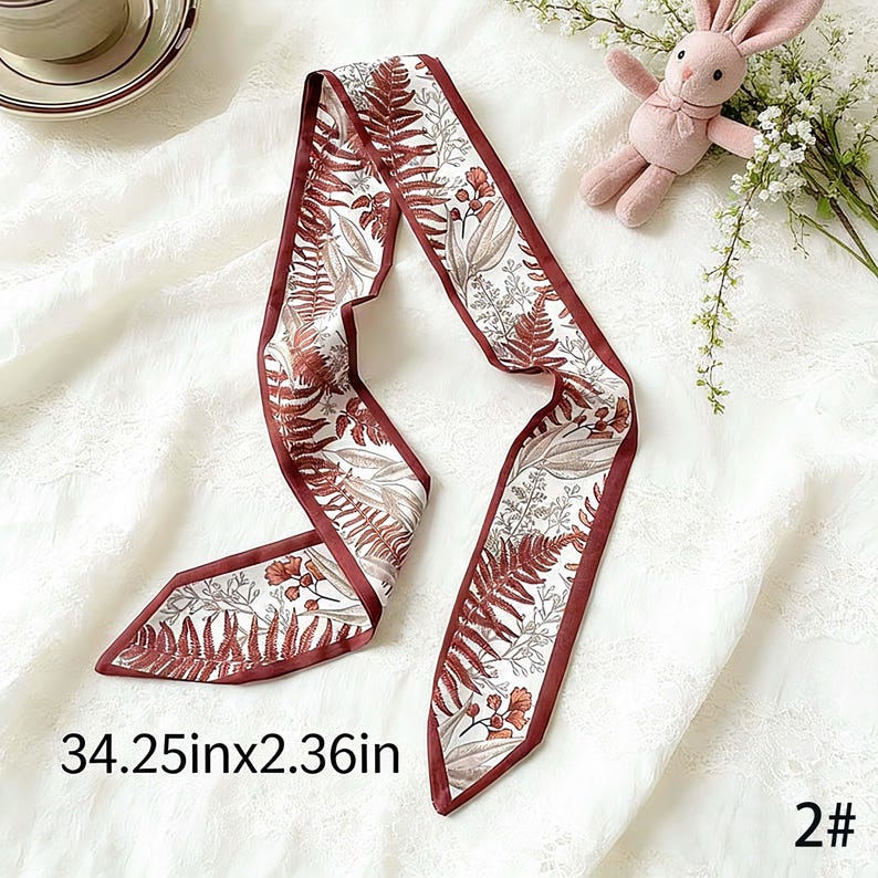 May include: A decorative scarf with a botanical print featuring ferns and flowers in shades of brown and white. The scarf has a dark red border and measures 34.25 inches long and 2.36 inches wide.