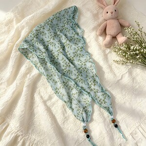 May include: A light blue triangular headscarf featuring a green leaf pattern, finished with a scalloped edge. The two bottom corners of the scarf are adorned with decorative beads in white, brown, and black, attached to thin fabric ties.
