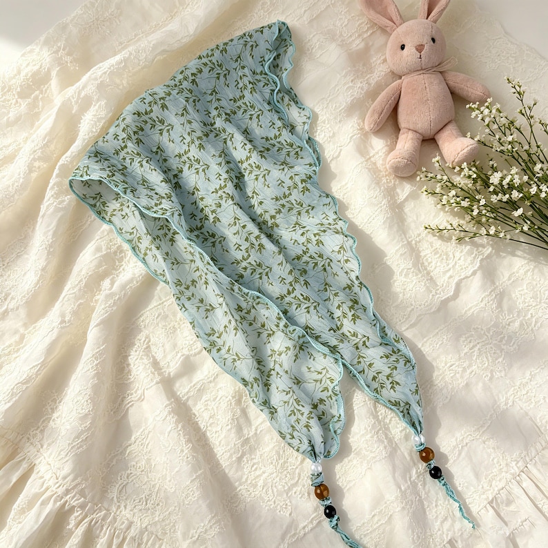 May include: A light blue triangular headscarf featuring a green leaf pattern, finished with a scalloped edge. The two bottom corners of the scarf are adorned with decorative beads in white, brown, and black, attached to thin fabric ties.