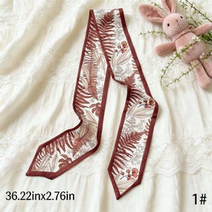 May include: A decorative scarf with a botanical print featuring ferns and flowers in shades of brown and beige on a white background. The scarf has a dark red border and measures 36.22 inches long and 2.76 inches wide.