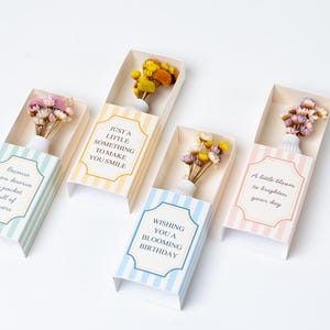 May include: Four decorative gift boxes with pastel stripes, each holding a small vase of dried flowers. The boxes have text that reads: "Because you deserve a pocket full of flowers," "Just a little something to make you smile," "Wishing you a blooming birthday," and "A little bloom to brighten your day."