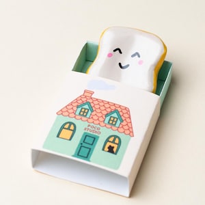 May include: A whimsical ceramic toast slice with a smiling face peeks out of a matchbox-style container. The container features a green house design with a red roof and the text "POCO STUDIO". The toast has yellow edges and is nestled in a light green interior.