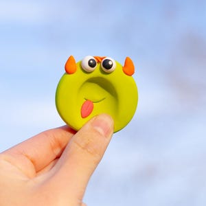 May include: A round, lime green clay charm featuring a cartoon monster face with two white eyes, orange horns, and a pink tongue sticking out. The surface has a smooth, matte texture with a small indentation in the center of the face.