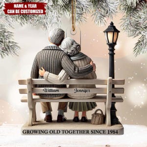 Puede incluir: Adorno decorativo que representa a una pareja de ancianos sentados en un banco del parque, abrazándose. El banco está inscrito con los nombres "William" y "Jennifer". La base dice "GROWING OLD TOGETHER SINCE 1984". También hay una farola y un bolso pequeño.