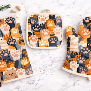 Peut inclure: Un ensemble de cuisine avec un motif de pattes de chat orange, beige et gris. L'ensemble comprend un torchon rectangulaire, un dessous de plat carré et une manique. Une cuillère en bois, une branche de romarin et des épices sont également présentes.