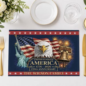 May include: A patriotic placemat featuring the American flag, a bald eagle, the Liberty Bell, and the Statue of Liberty. The text reads "United States of America 1776-2026 250th Anniversary" and "The Wilson Family". Gold flatware and a white plate are also visible.