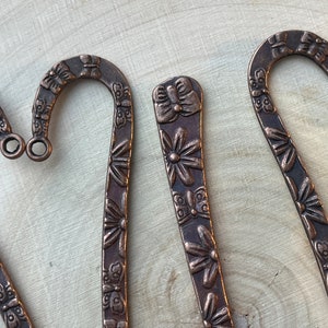 May include: Three copper bookmark pendants with a floral and butterfly design. The bookmarks are shaped like a hook and have a decorative design.
