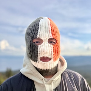 Op de afbeelding: Een gebreide bivakmuts in een driekleurig ontwerp van grijs, wit en oranje. Het skimasker bedekt het hele hoofd en de nek, met openingen voor de ogen en mond. De persoon draagt een lichtgroene hoodie en een zwart-grijze jas.