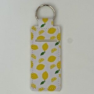 May include: A white keychain holder with a pattern of yellow lemons and green leaves. The holder has a silver key ring and a rectangular opening. The design features whole and sliced lemons.
