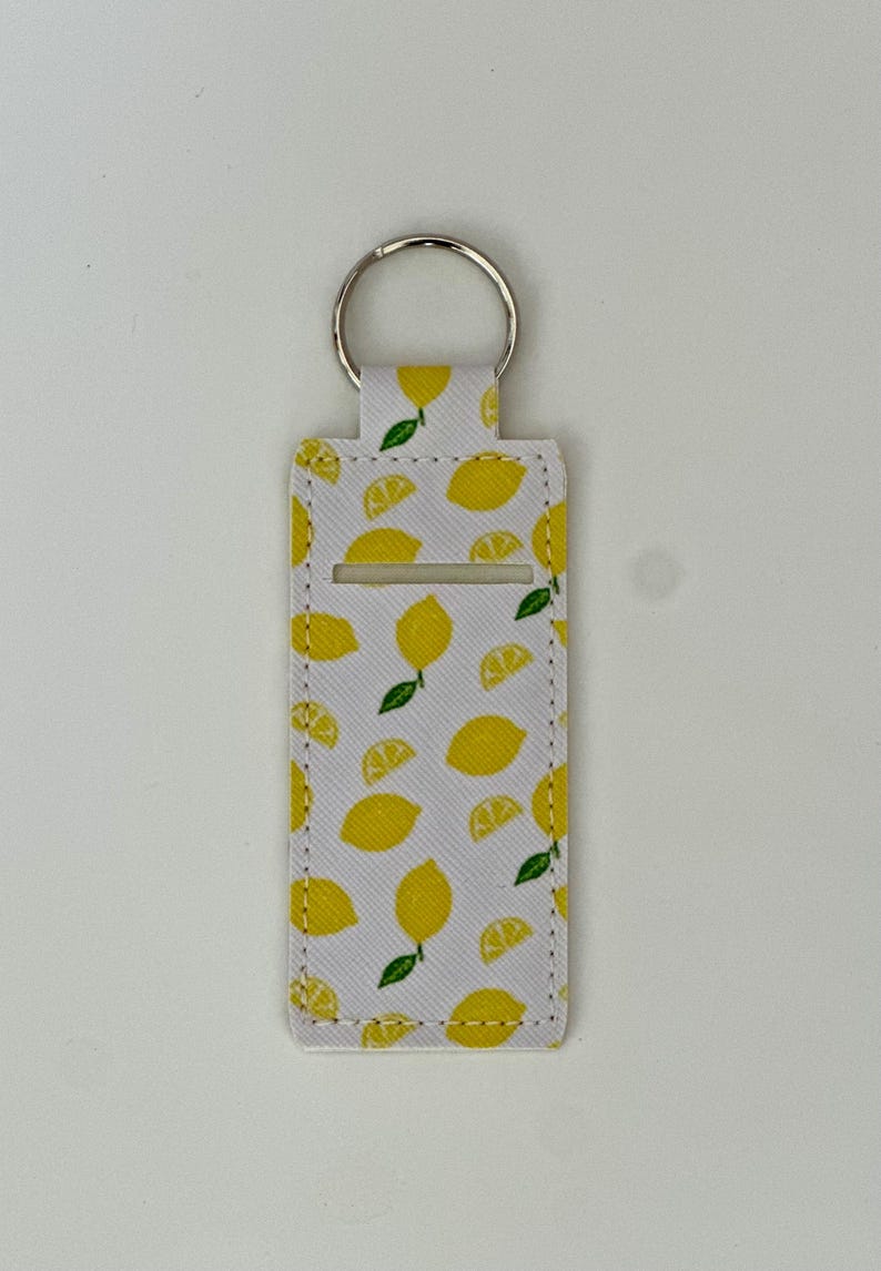 May include: A white keychain holder with a pattern of yellow lemons and green leaves. The holder has a silver key ring and a rectangular opening. The design features whole and sliced lemons.