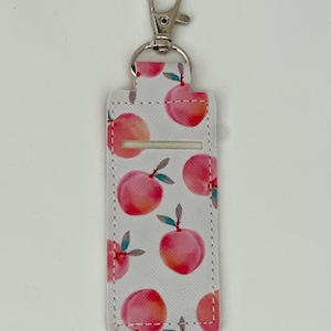 May include: A white lip balm holder with a silver clip, patterned with watercolor-style peaches. The peaches are pink and orange with green leaves. The holder has a rectangular shape with a slot for the lip balm.