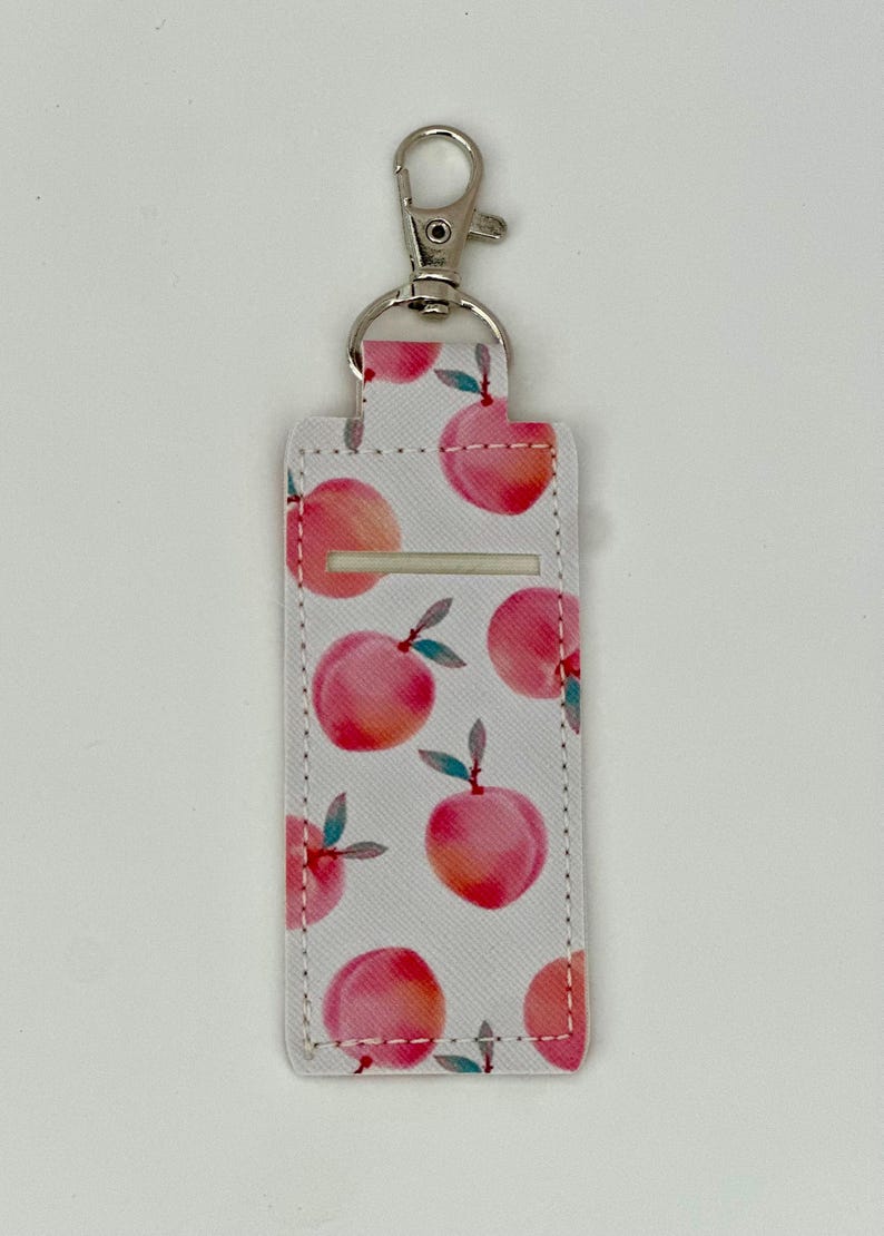 May include: A white lip balm holder with a silver clip, patterned with watercolor-style peaches. The peaches are pink and orange with green leaves. The holder has a rectangular shape with a slot for the lip balm.