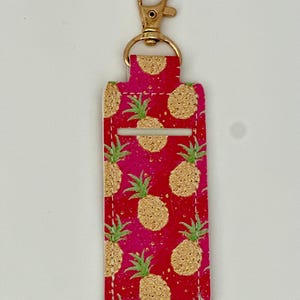 May include: A red and pink lip balm holder with a gold-colored clasp. The holder is decorated with a repeating pattern of gold pineapples with green leaves. The holder is rectangular with a slot for the lip balm.