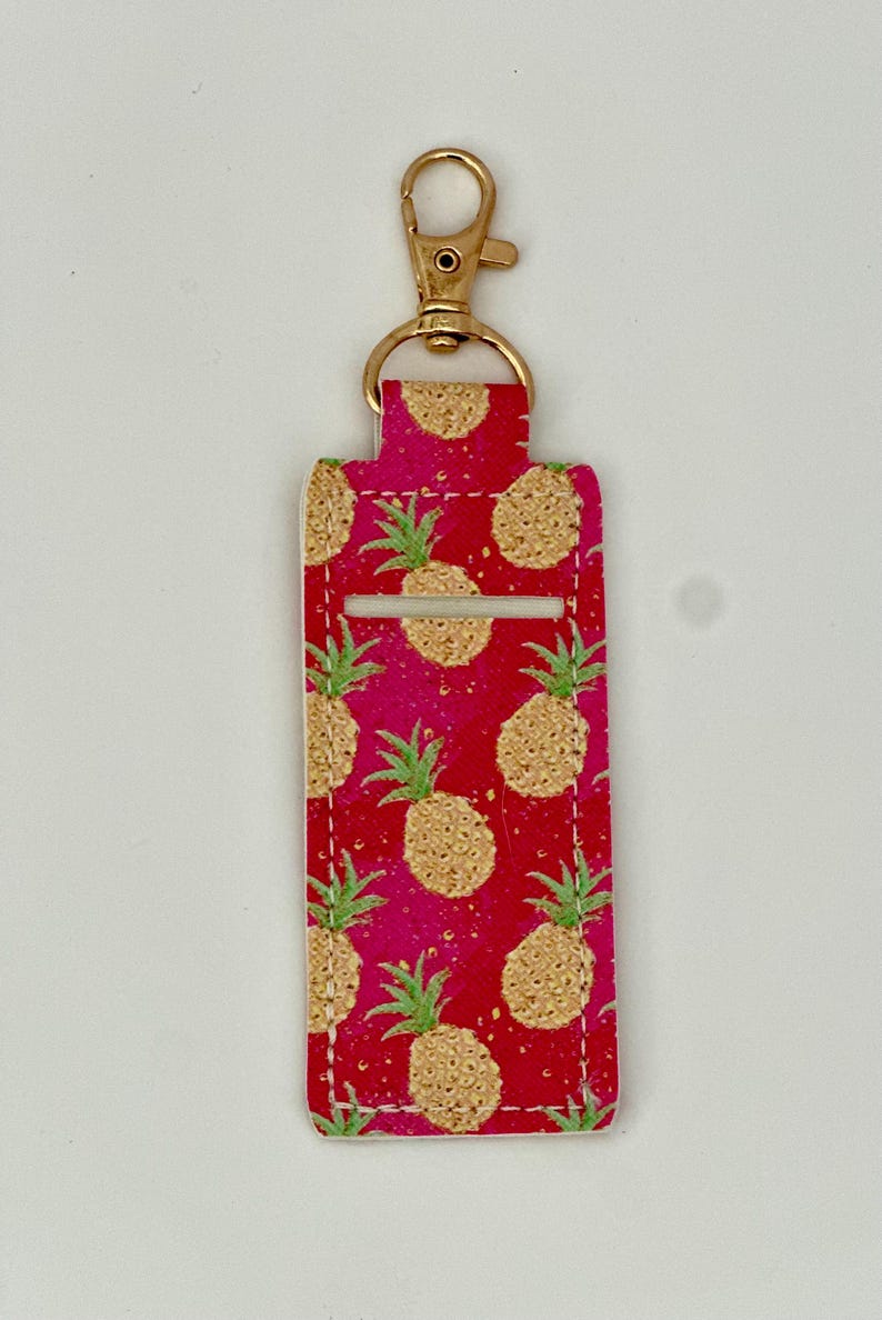 May include: A red and pink lip balm holder with a gold-colored clasp. The holder is decorated with a repeating pattern of gold pineapples with green leaves. The holder is rectangular with a slot for the lip balm.
