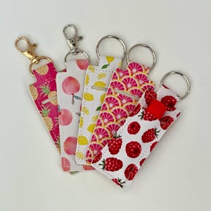 May include: Five lip balm holders with keyrings. The holders feature various fruit patterns, including pineapples, peaches, lemons, pink citrus slices, and raspberries. Each holder has a different coloured keyring.