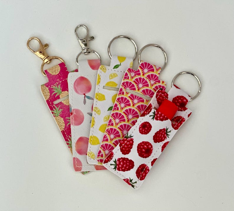 May include: Five lip balm holders with keychains. The holders feature various fruit patterns, including pineapples, peaches, lemons, pink citrus slices, and raspberries. Each holder has a different colored keychain.