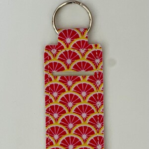May include: A rectangular keychain holder with a red and yellow citrus fruit pattern. The holder has a silver key ring attached to the top. The design features a repeating pattern of red citrus slices with yellow details.