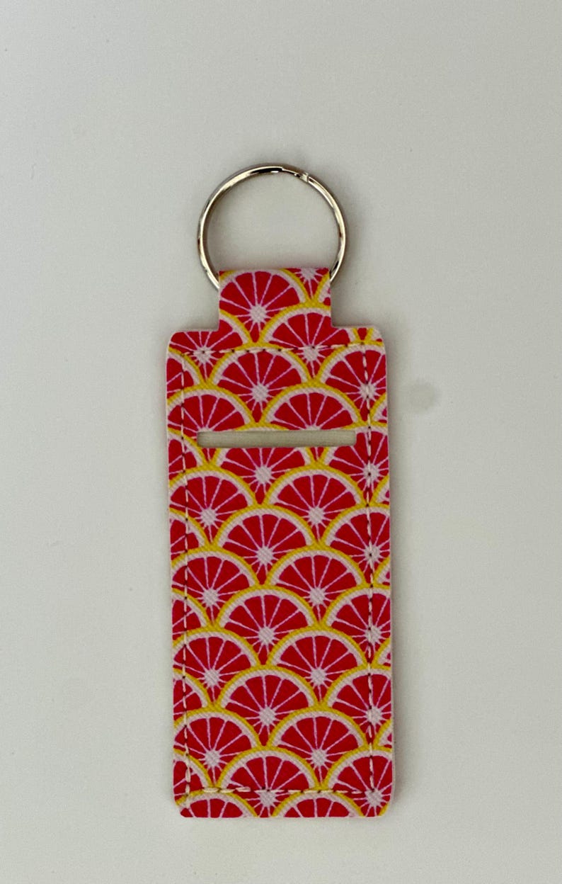 May include: A rectangular keychain holder with a red and yellow citrus fruit pattern. The holder has a silver key ring attached to the top. The design features a repeating pattern of red citrus slices with yellow details.