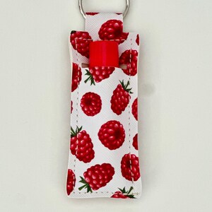 May include: A white lip balm holder with a pattern of red raspberries and green leaves. A silver key ring is attached to the top. A red lip balm is inserted in the holder.
