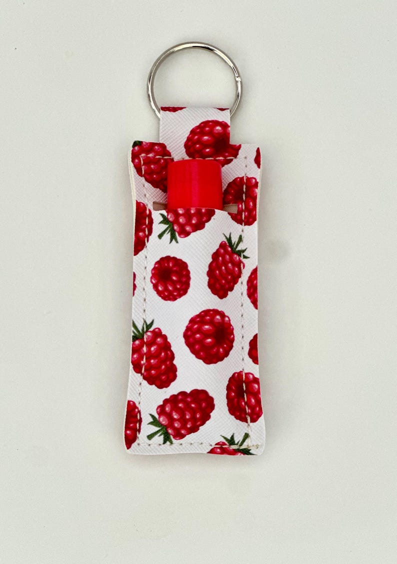 May include: A white lip balm holder with a pattern of red raspberries and green leaves. A silver key ring is attached to the top. A red lip balm is inserted in the holder.
